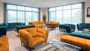 A spacious room with teal and mustard upholstered sofas, a glass coffee table, and large windows with sheer curtains. - Glass House Retreat (Upminster)