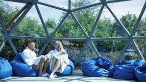 A couple sitting on blue bean bags under a geometric glass structure with greenery in the background. - Glass House Retreat (Upminster)