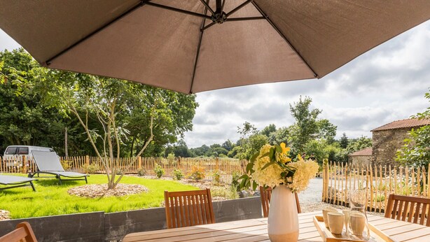 A wooden table with a vase of flowers and a tray with glasses, under a large umbrella, in a garden setting with a fence and trees.