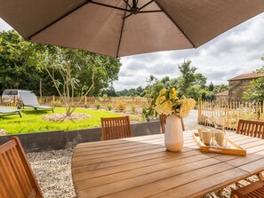 A wooden table with a vase of flowers and a tray with glasses, under a large umbrella, in a garden setting with a fence and trees.