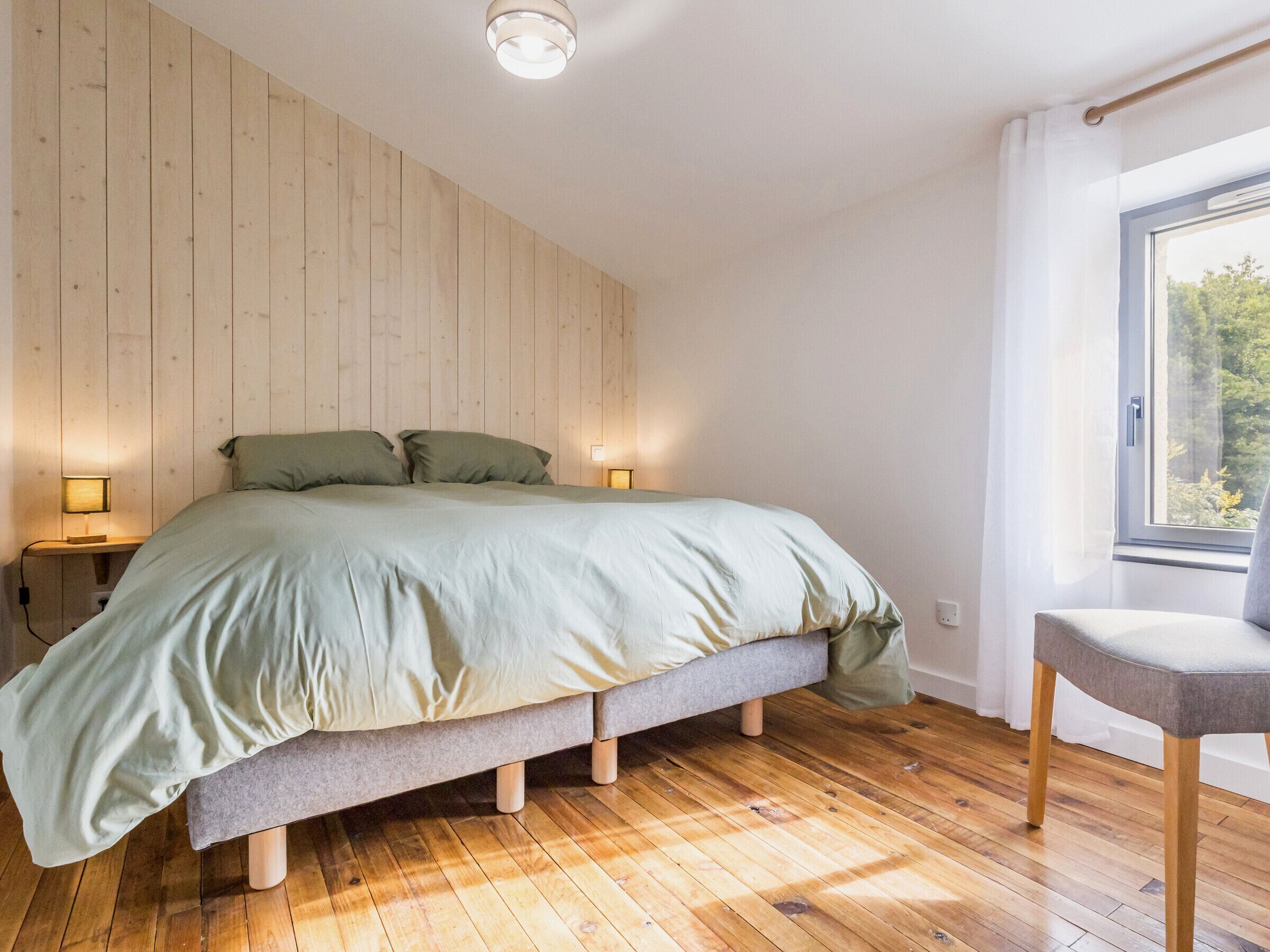 A bedroom with a wooden floor, a bed with a grey mattress, two bedside lamps, and a window with a view of trees.