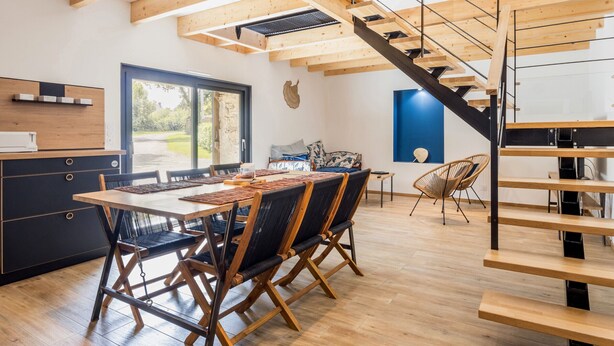 A modern kitchen with wooden beams, a dining area with a large table and chairs, and a staircase with a glass railing.