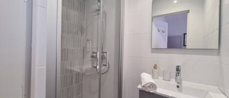 Un baño moderno con ducha de vidrio, un lavabo blanco con un espejo encima y un cubo de basura pequeño.