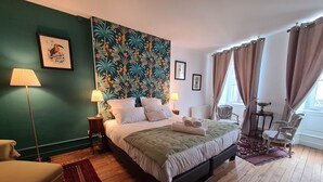 A bedroom with a large bed, two bedside tables, a chair, a window with curtains, and a tropical-themed wall mural. - Spacieuse Chambre avec Lit King-Size, Salle d'Eau Privative et Petit-Déjeuner Inclus (Châteauroux)