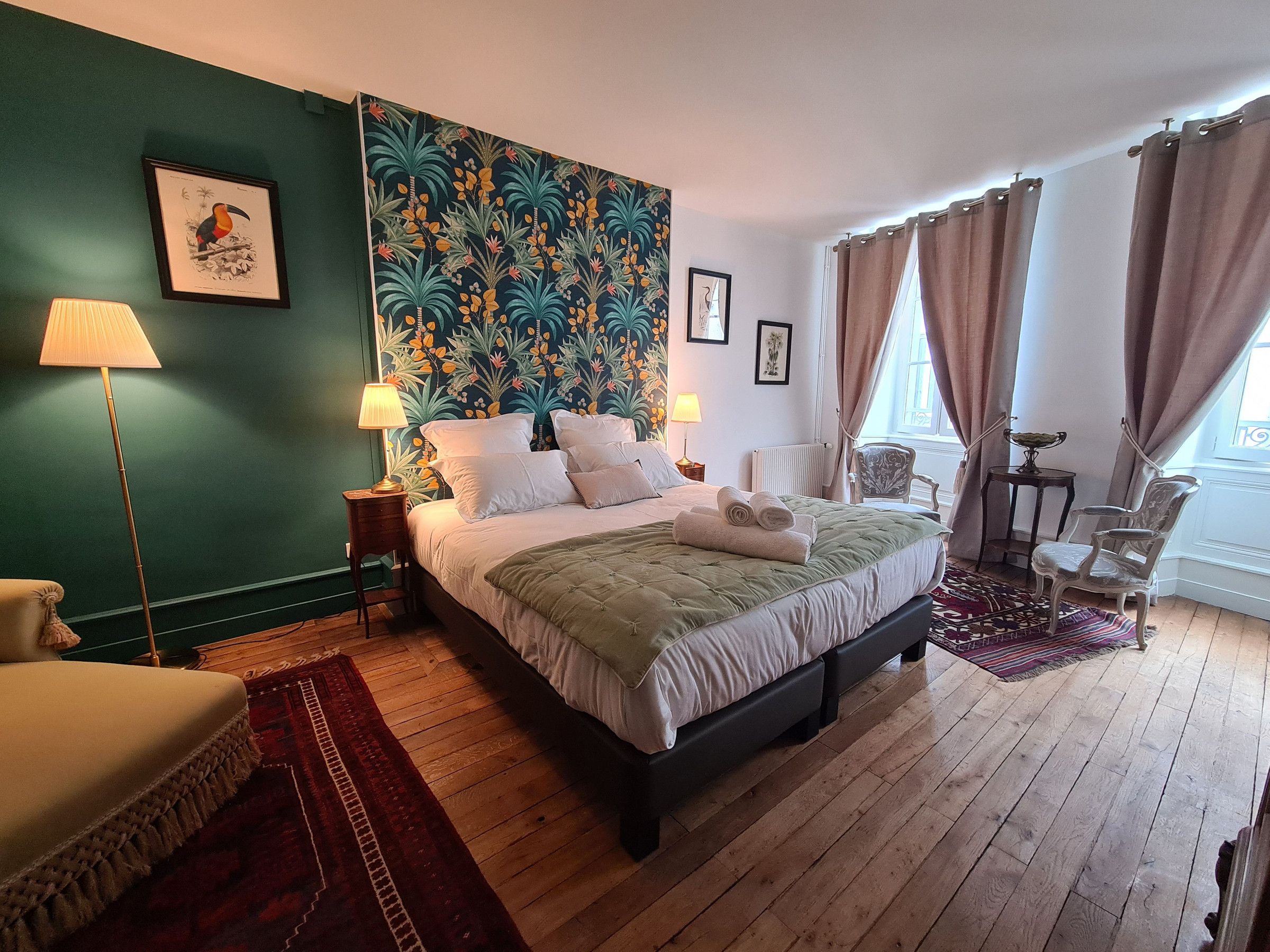Ein Schlafzimmer mit einem großen Bett, zwei Nachttischen, einem Sessel, einem Fenster mit Vorhängen und einem Wandbild im tropischen Stil.