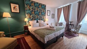 Ein Schlafzimmer mit einem großen Bett, zwei Nachttischen, einem Sessel, einem Fenster mit Vorhängen und einem Wandbild im tropischen Stil.