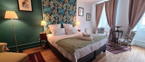 A bedroom with a large bed, two bedside lamps, a headboard with a tropical print, a chair, a small table, and a window with curtains.