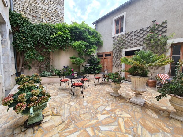 Une cour avec un sol en pierre, une table entourée de chaises, des plantes en pot et un mur de pierre orné de plantes grimpantes.