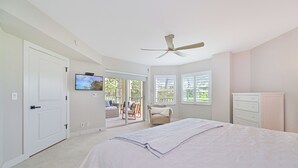 Un dormitorio con una cama, ventilador de techo, televisor y puerta corrediza de vidrio que da a un balcón.