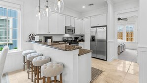 Una cocina moderna con armarios blancos, encimeras de granito y electrodomésticos de acero inoxidable. Hay taburetes de bar con asientos acolchados y luces colgantes sobre la isla.