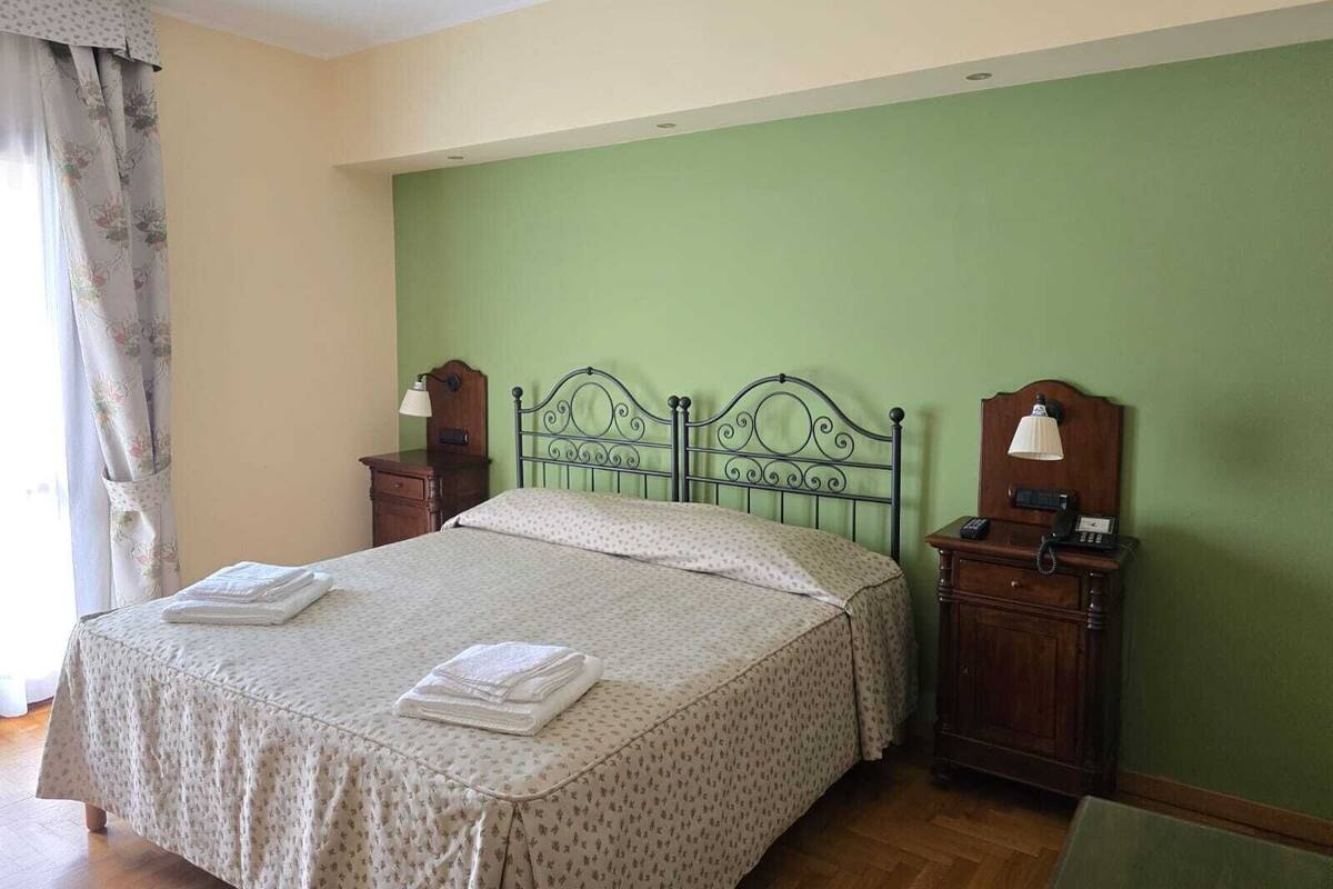 A neatly made bed with a patterned bedspread and two folded towels, a nightstand with a lamp, and a wooden dresser with a telephone.