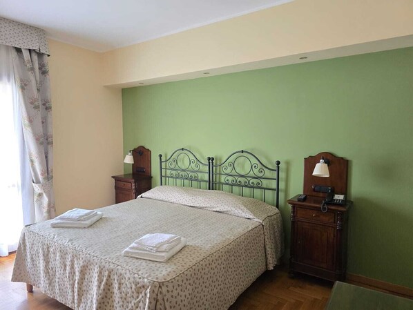 Una cama bien hecha con una colcha estampada y dos toallas dobladas, una mesita de noche con una lámpara, y un aparador de madera con un teléfono.
