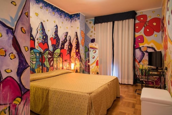A hotel room with a bed, a desk, and a chair. The walls are decorated with colorful artwork.
