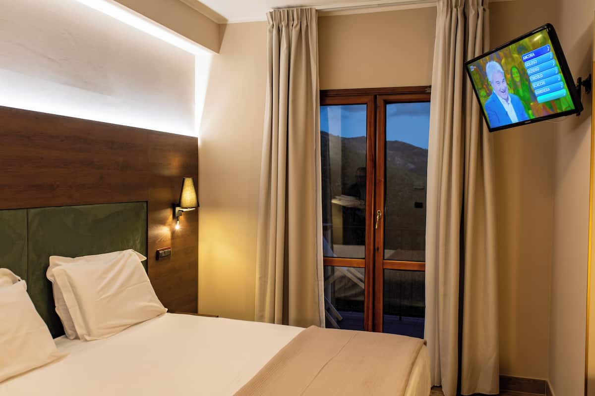 A hotel room with a large bed, a headboard, a wall-mounted TV, and a window with curtains.