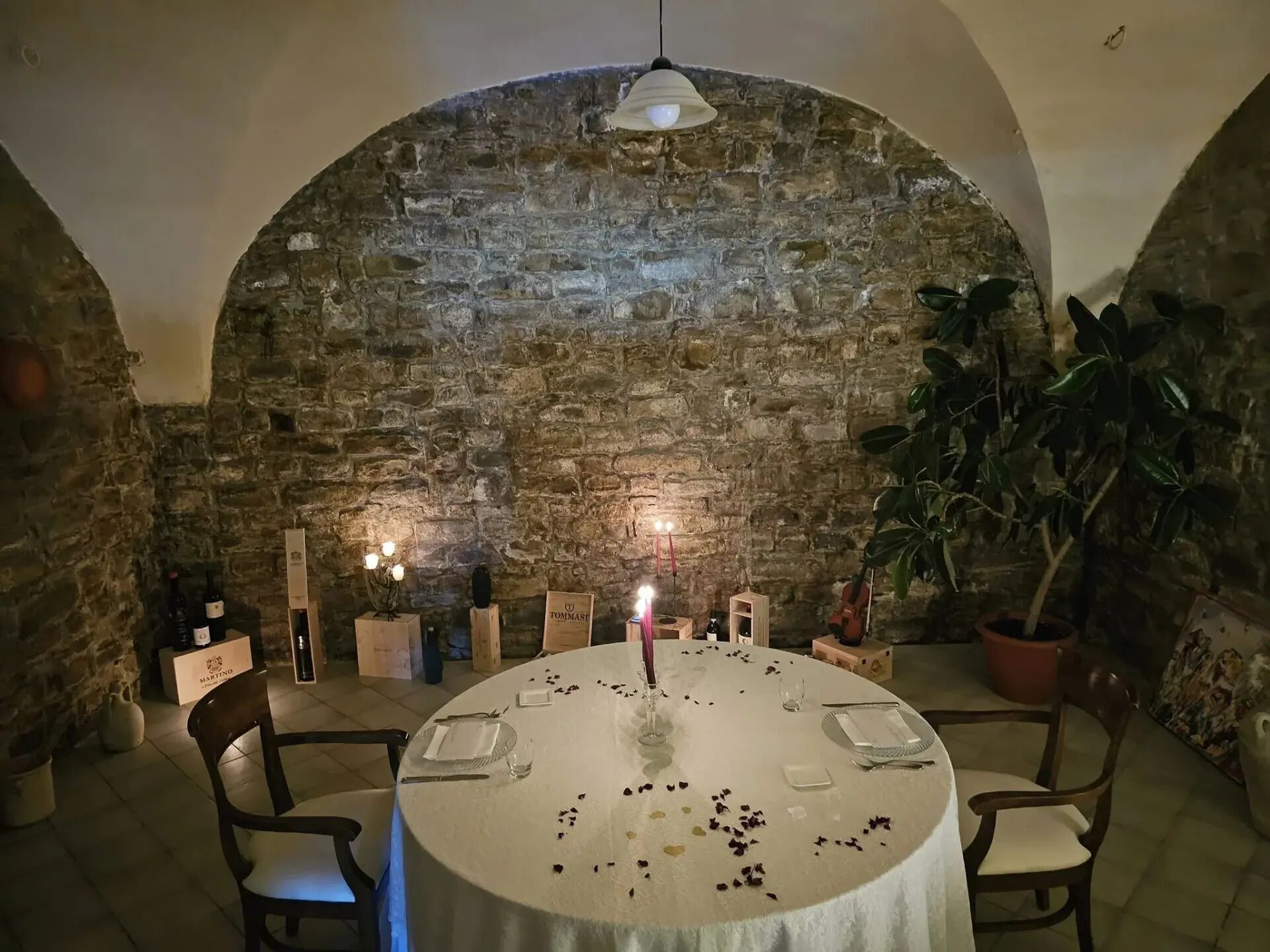 Uma mesa redonda com toalha branca, rodeada por cadeiras, num espaço com parede de pedra e teto em arco.