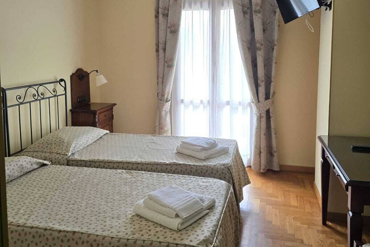 A room with two beds, a desk, a flat-screen TV mounted on the wall, and a ceiling light fixture.