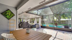 A covered patio with a wooden table and chairs, a grill, and a view of a swimming pool. - Screened Lanai + Fenced Yard: Luxe Orlando Home (Orlando)