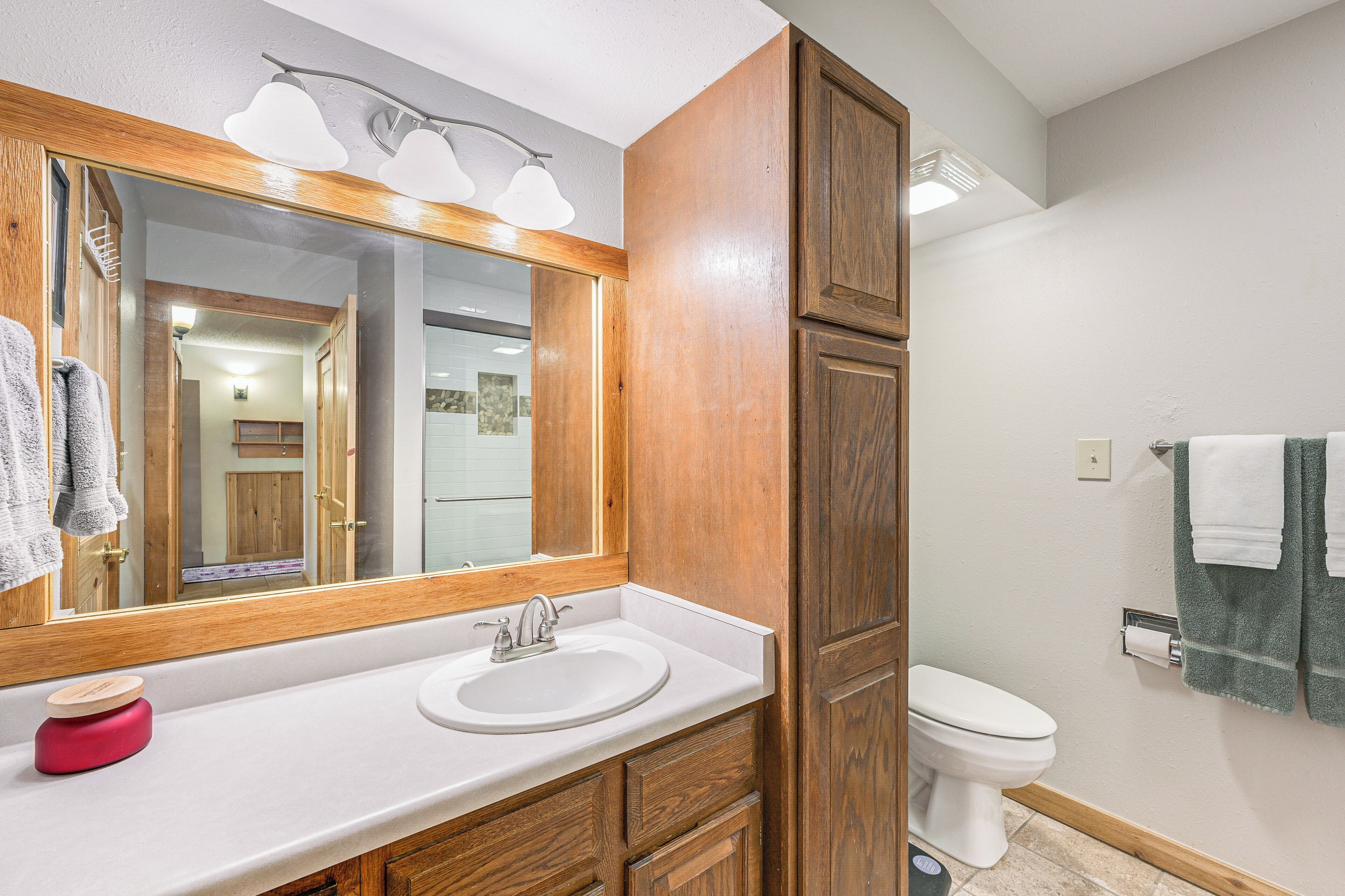 A bathroom with a wooden vanity, a white sink, a mirror with lights, a toilet, and a towel rack.