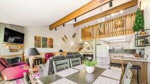 Een moderne woonkamer met een eethoek, houten balken, een televisie en een trap naar een hoger gelegen verdieping.