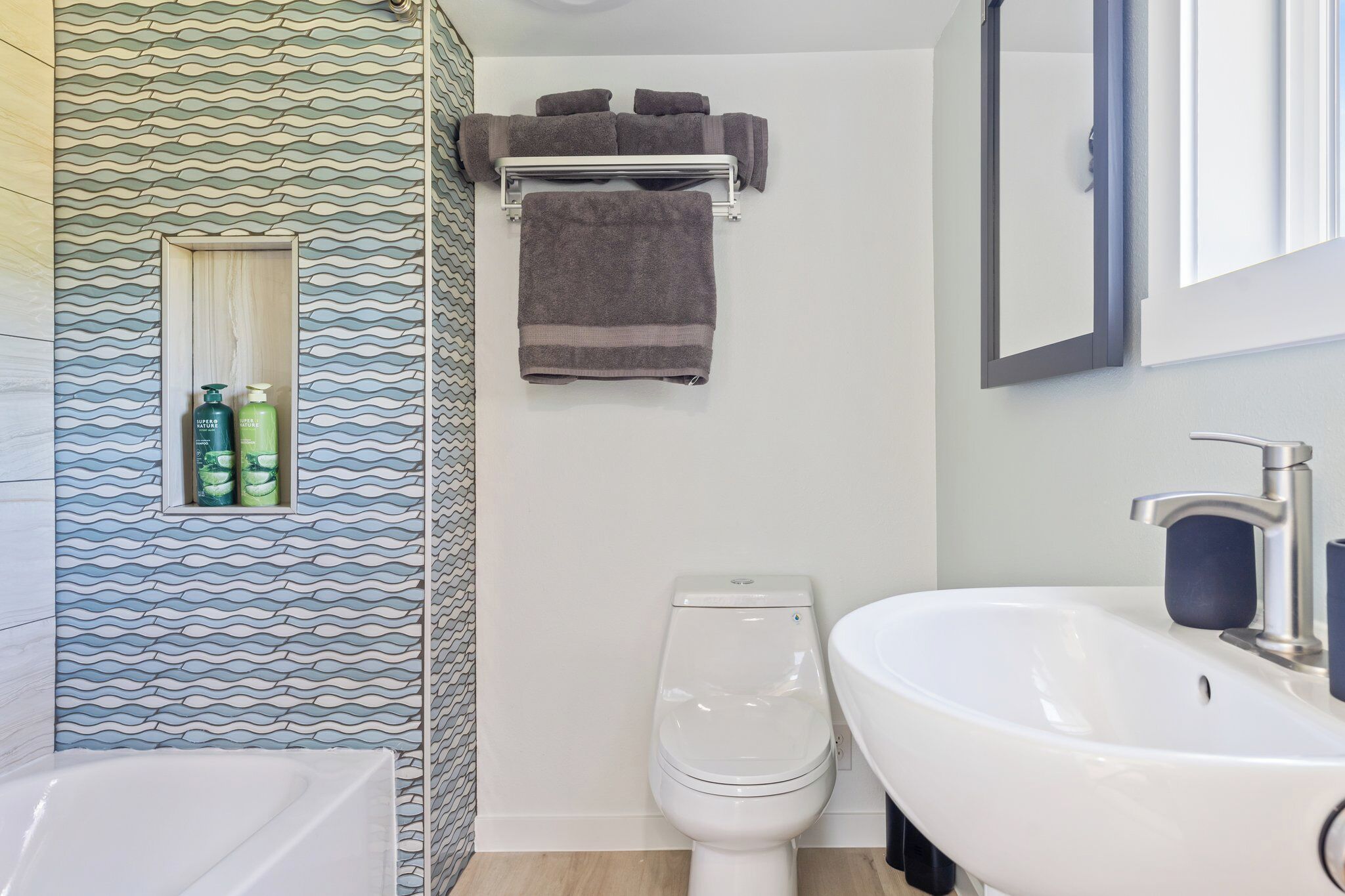Un baño con un lavabo blanco, un inodoro y un perchero con toallas. Hay un estante pequeño con artículos de aseo.