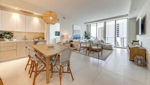 A modern kitchen with white cabinets, a dining area with a wooden table and wicker chairs, and a living room with a sofa and large windows. - Ocean Views & Pool Access: Riviera Beach Condo! (Singer Island)