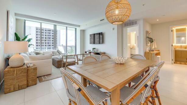A modern living room with a wooden dining table, wicker furniture, and a large window offering a city view. - Ocean Views & Pool Access: Riviera Beach Condo! (Singer Island)