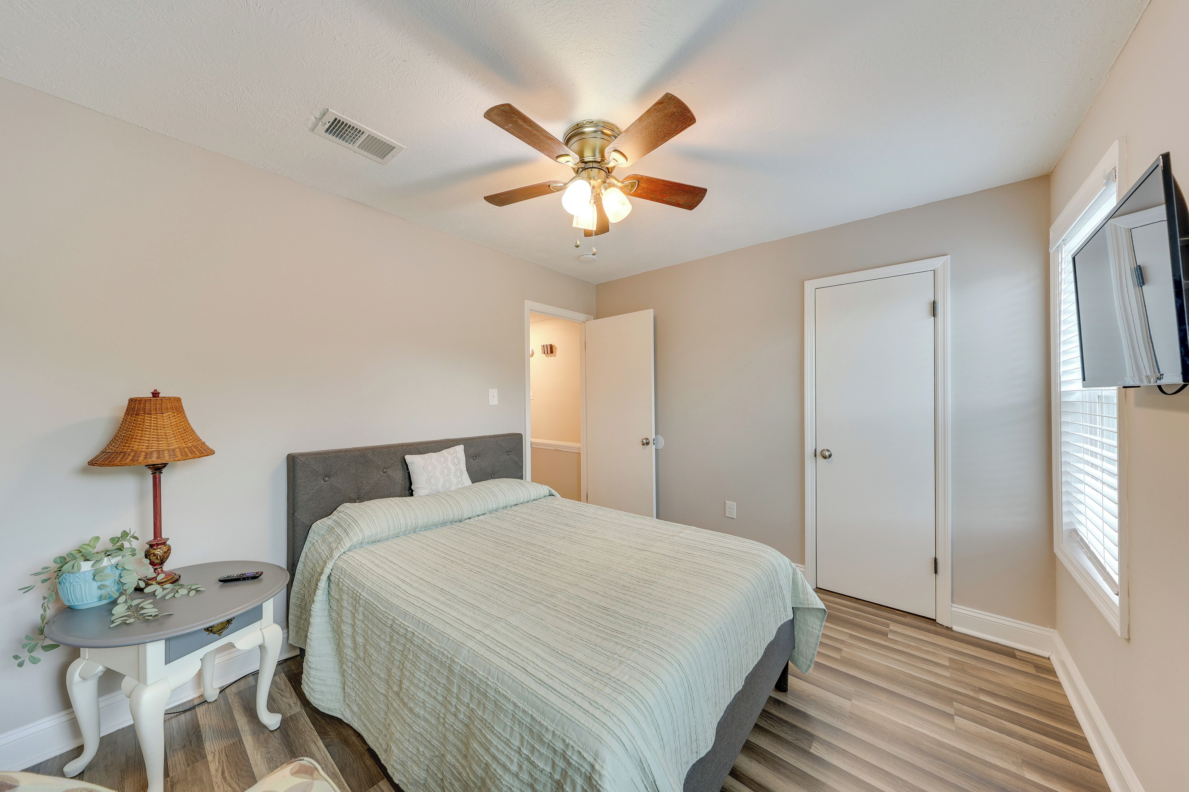 A bedroom with a bed, a ceiling fan, a bedside table with a lamp, and a television on the wall.
