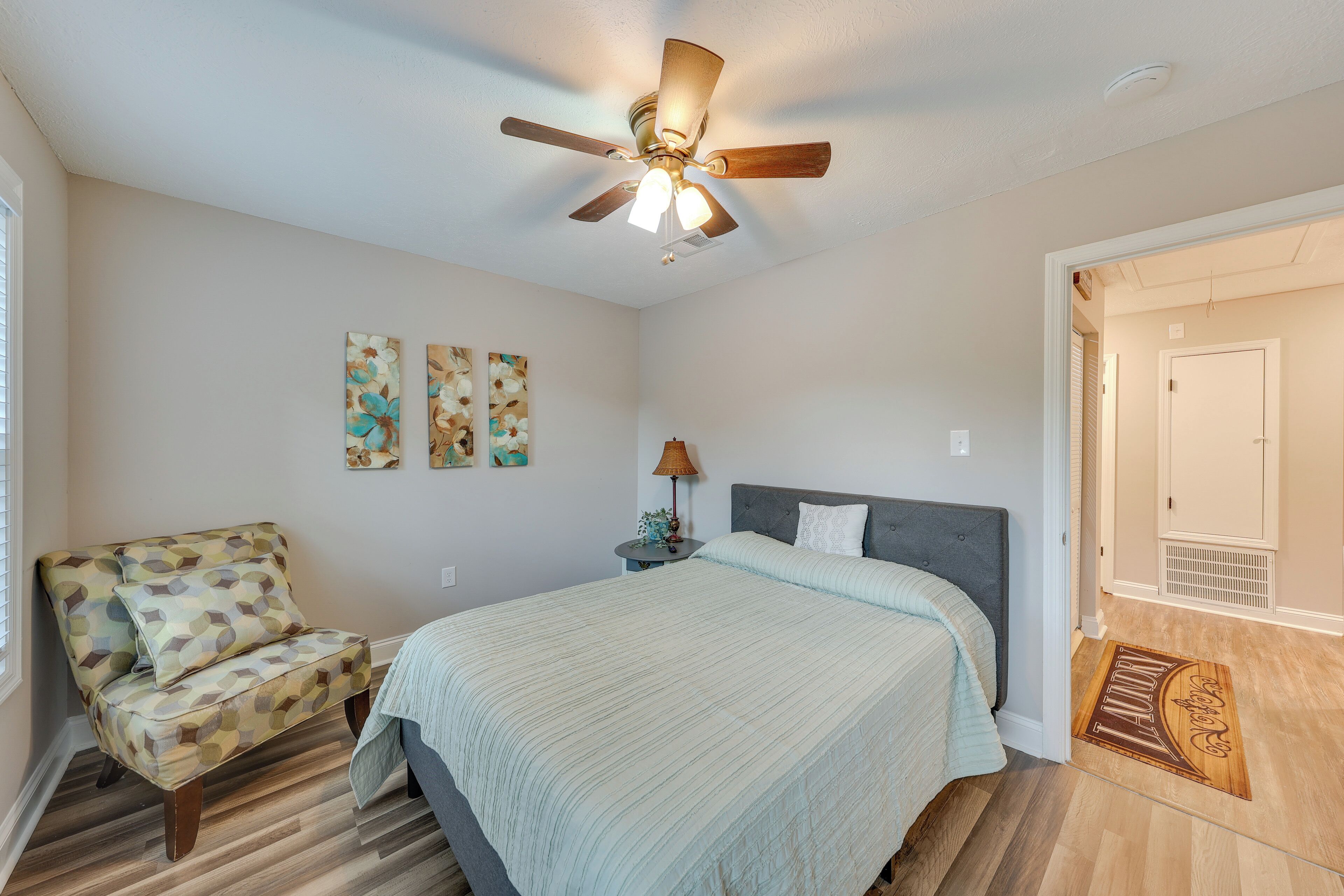 A bedroom with a bed, a chair, a ceiling fan, and artwork on the wall.