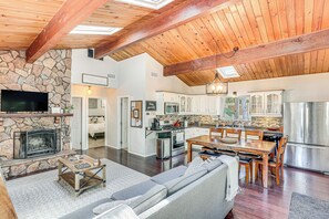 A spacious living area with a stone fireplace, a large sofa, a dining table with chairs, and a kitchen with modern appliances. - 1 Mi to Lake: Crestline Cabin w/ Deck + Gas Grill! (Crestline)