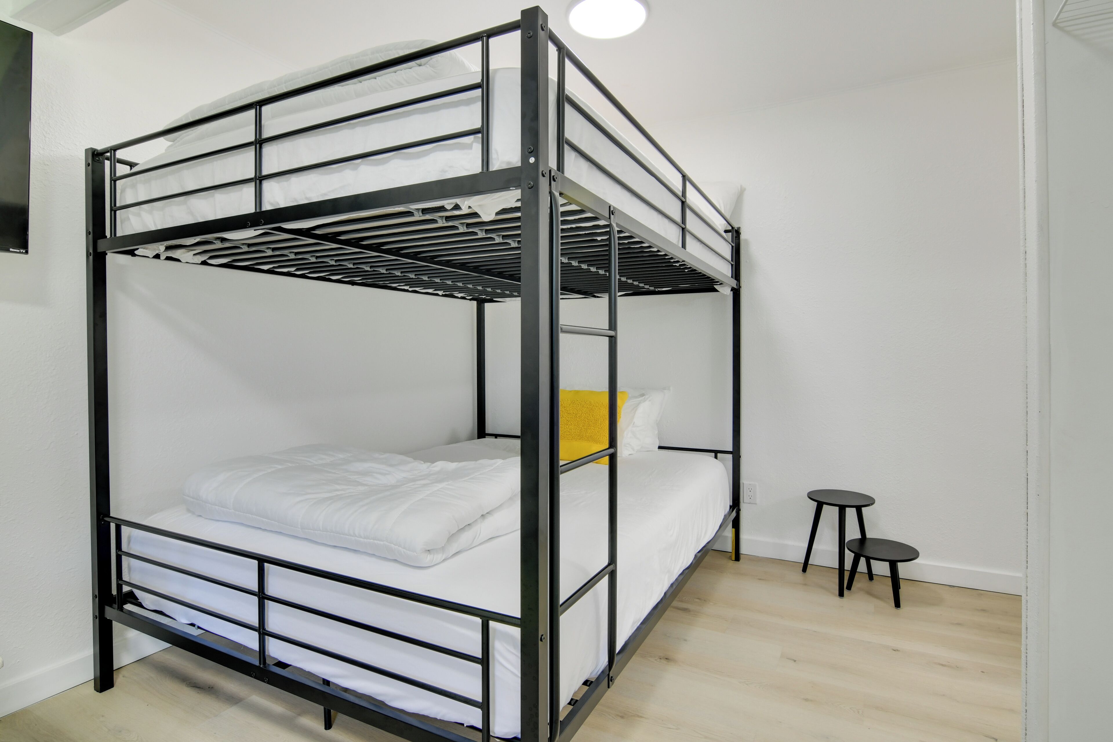 Una cama litera con ropa de cama blanca y una almohada amarilla, una mesita negra pequeña y una pared blanca con un interruptor de luz.