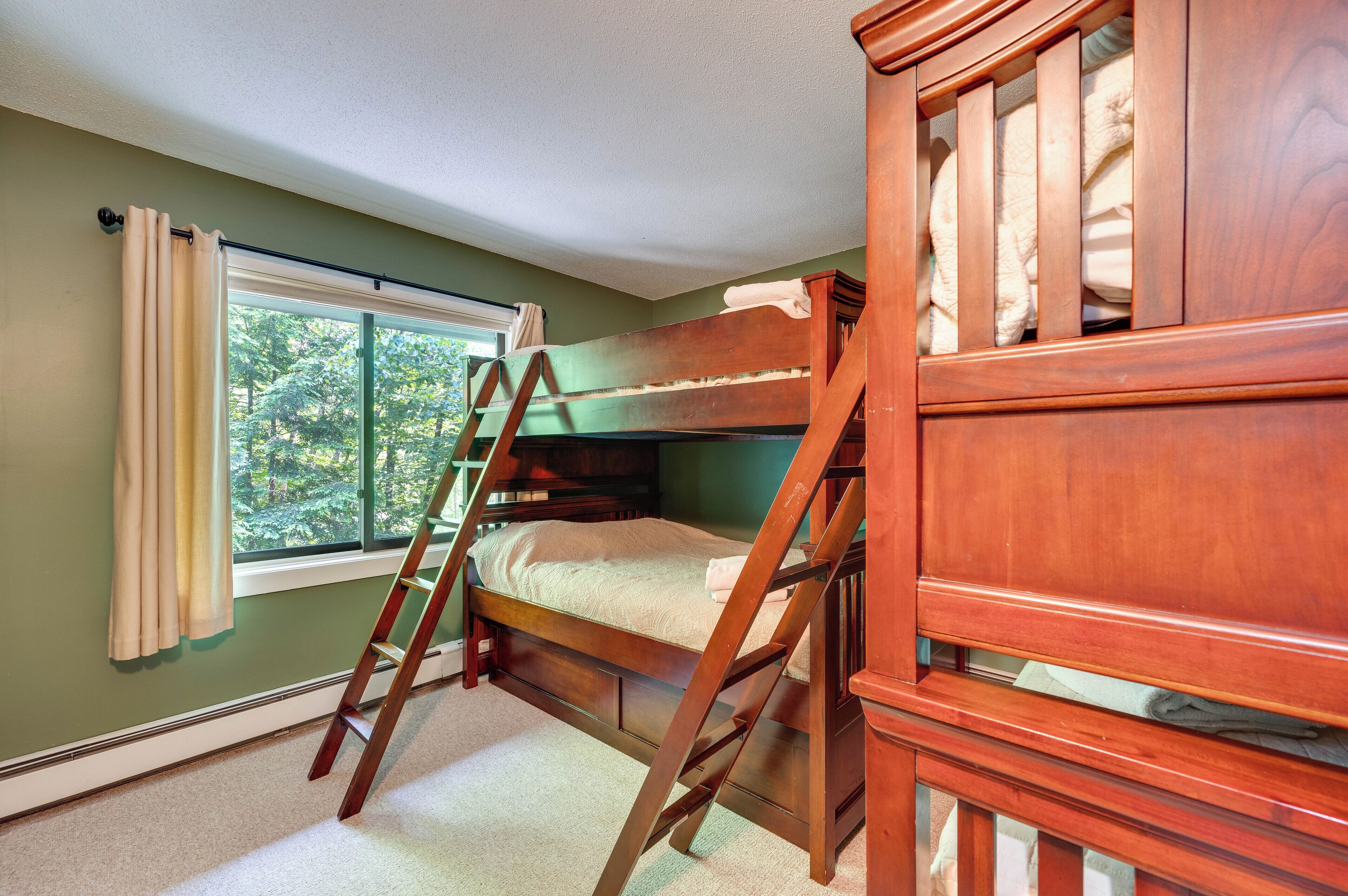 A bunk bed with a wooden ladder, a window with curtains, and a view of trees outside.