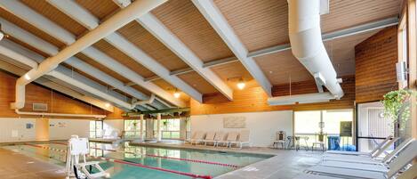 An indoor swimming pool with a wooden ceiling, clear ceiling pipes, and lounge chairs along the poolside.