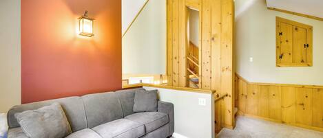 Una habitación con un sofá gris, una escalera de madera y una lámpara fijada a la pared.