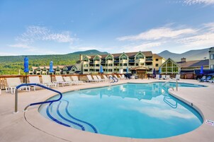 Piscina de resort con sillas de descanso y sombrillas, rodeada por un edificio con varios balcones y un paisaje montañoso de fondo.