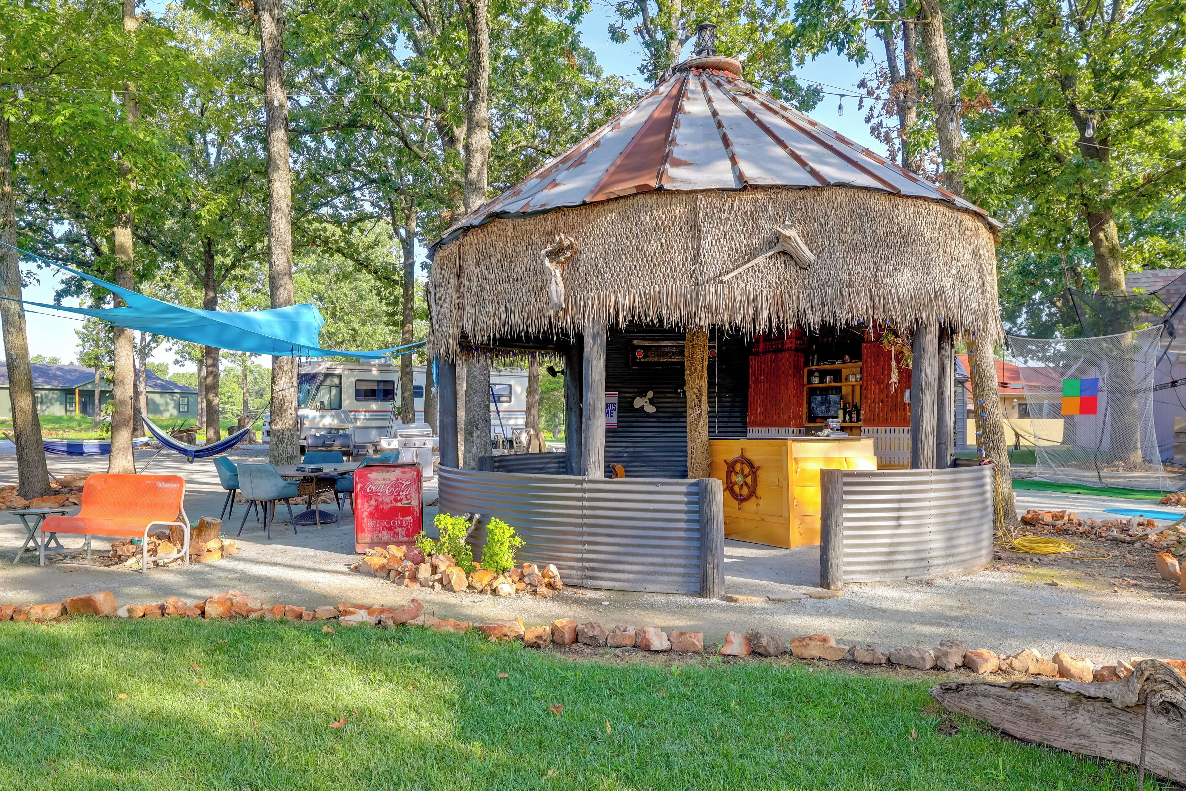 A rustic gazebo with a thatched roof, surrounded by trees and a small seating area with benches.