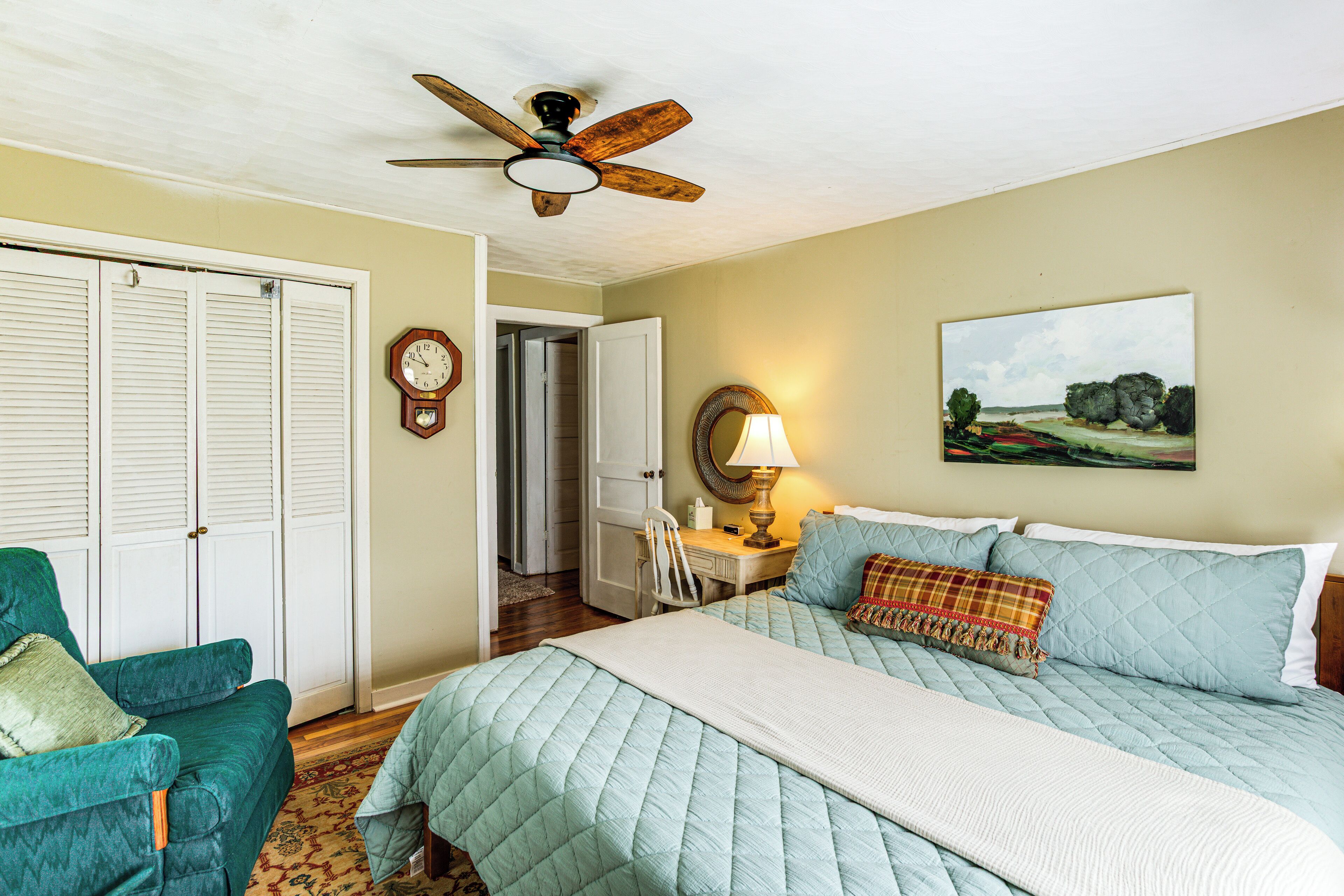 A bedroom with a bed, a chair, a ceiling fan, a clock, and a painting on the wall.