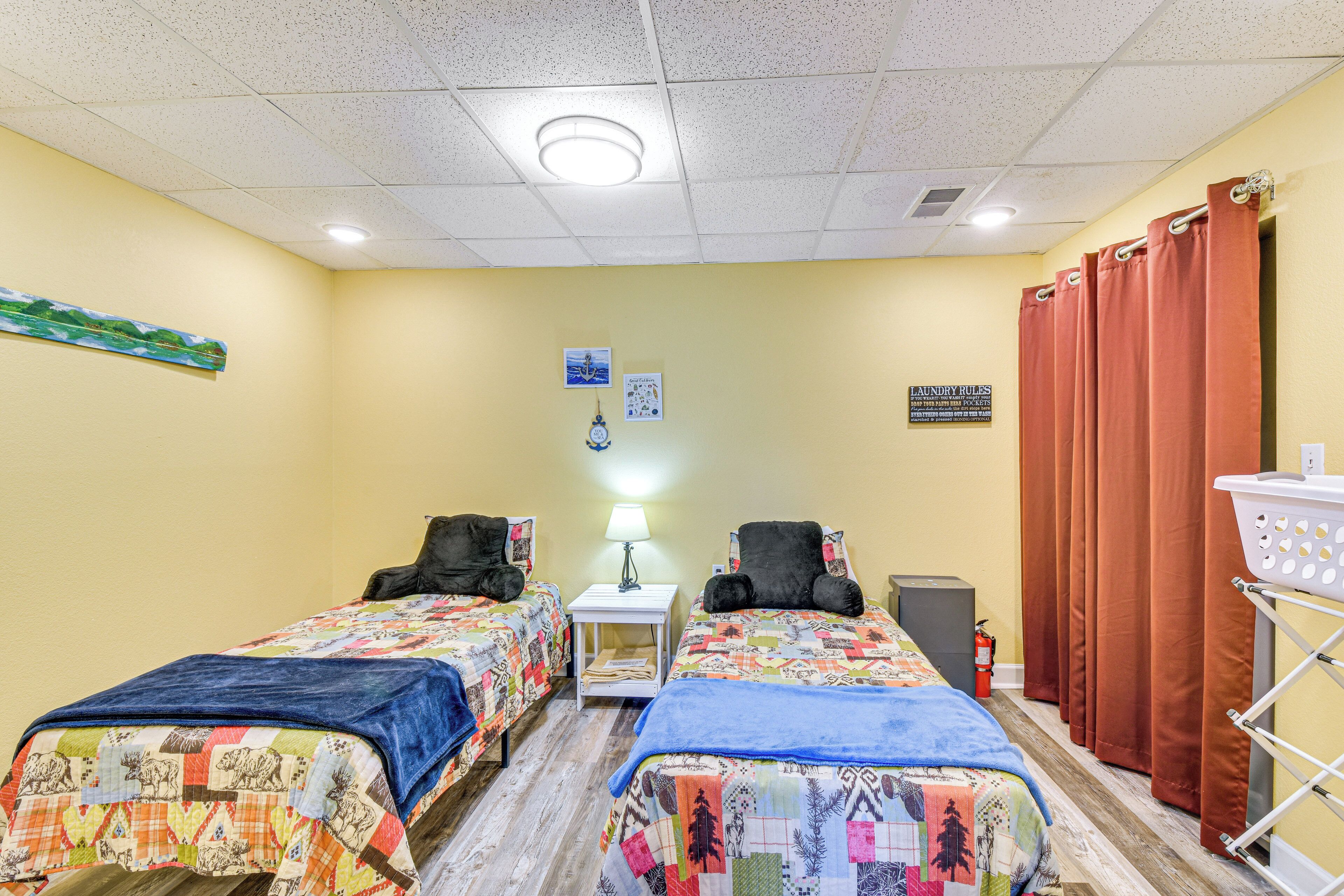 A hotel room with two beds, a small table, a lamp, a fire extinguisher, and a laundry cart.