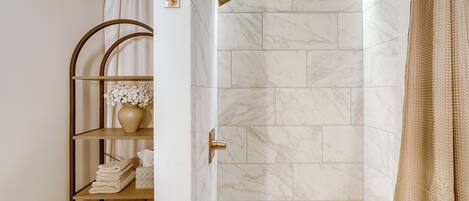 A bathroom with a shower, marble tiles, a towel rack, and a small wooden stool.