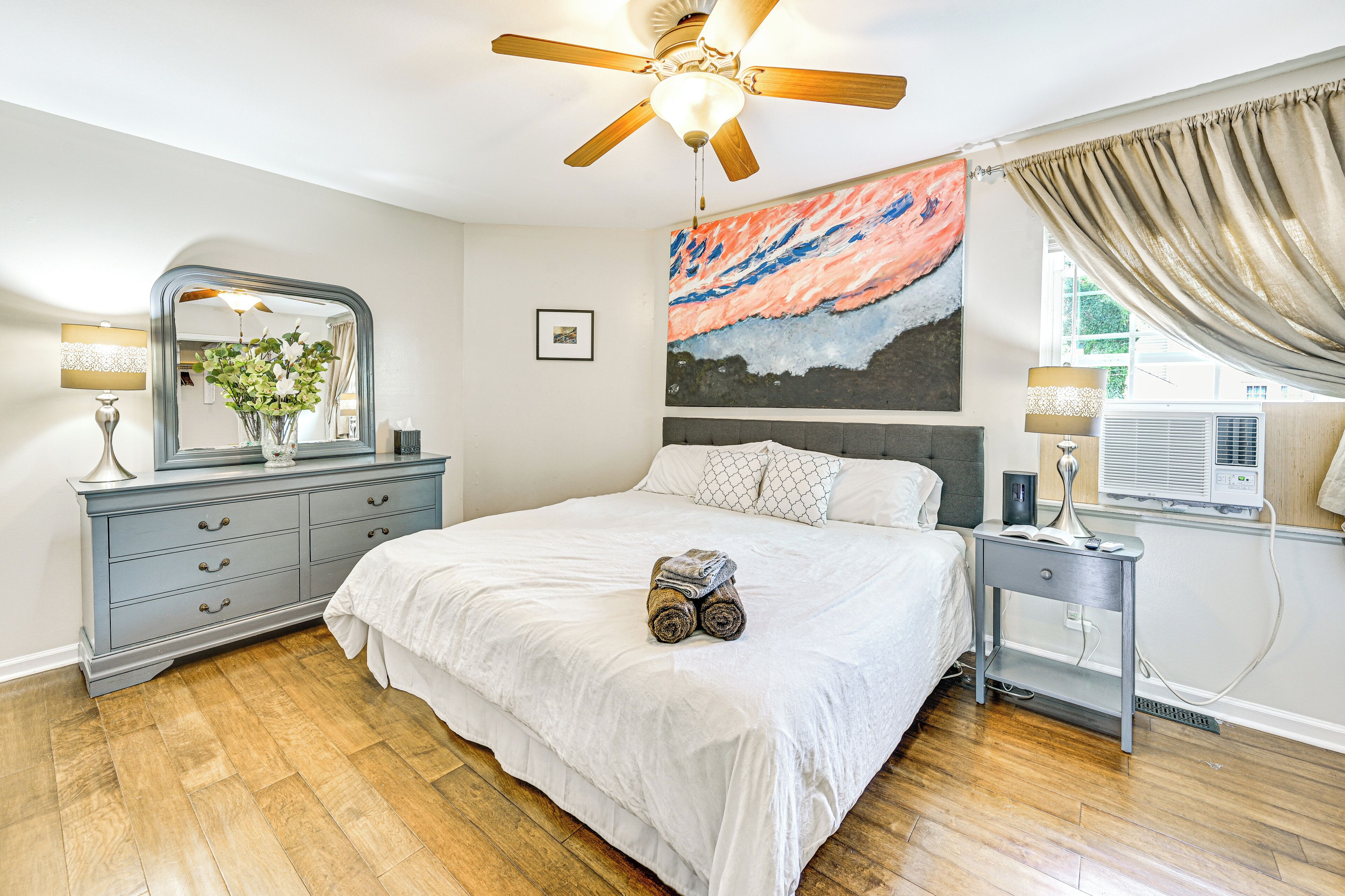 A bedroom with a bed, a dresser, a nightstand, a ceiling fan, a painting, and an air conditioning unit.