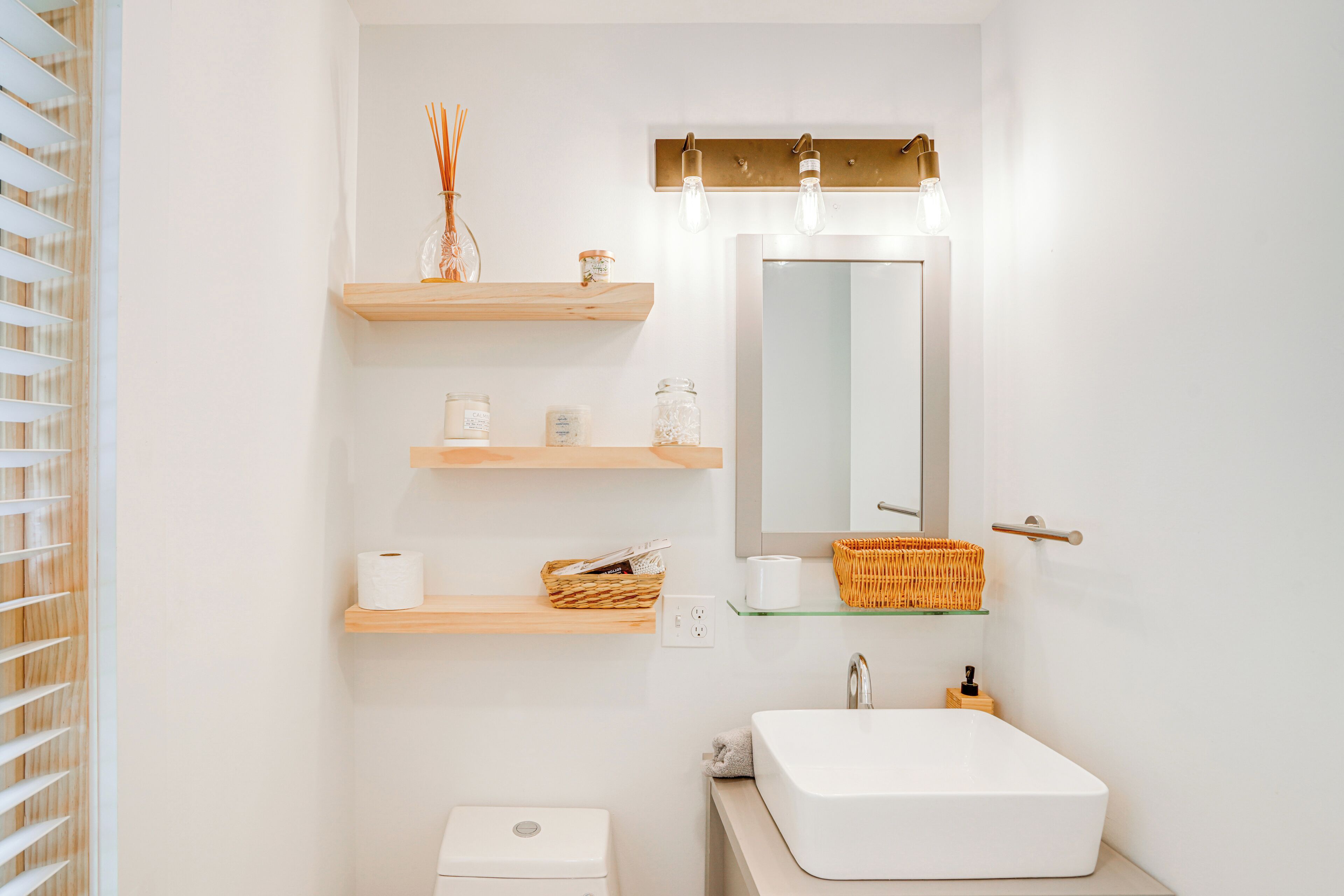 Une salle de bain moderne avec un lavabo blanc, un miroir et des étagères en bois où sont rangés les produits de toilette.