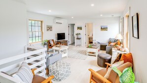 Un salón amplio con sofá, reposapiés y mesa de centro, televisor y una escalera que sube a otro nivel.