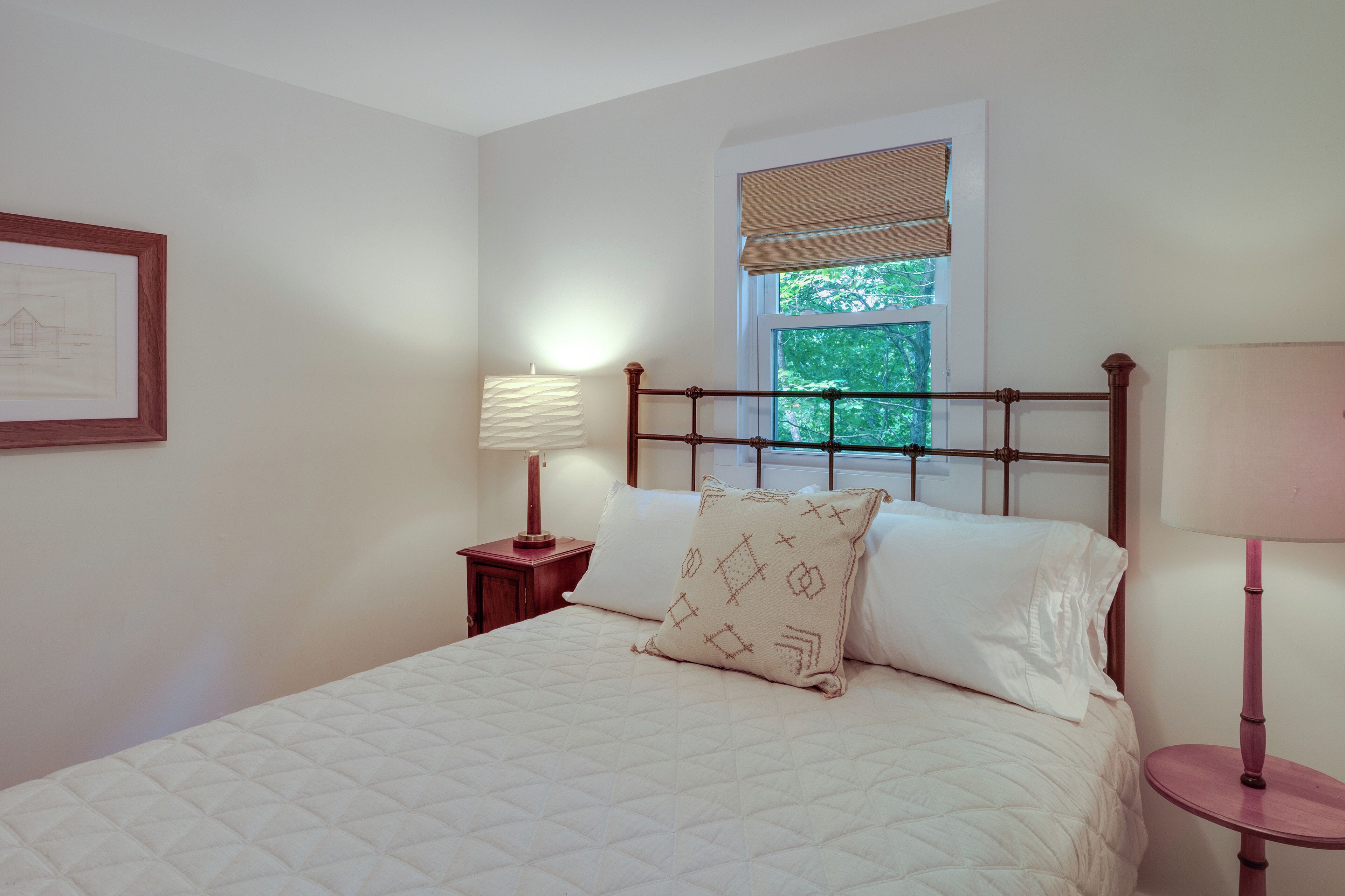 A neatly made bed with a quilted cover and two pillows, a bedside table with a lamp, a framed picture on the wall, and a window with a view of greenery.
