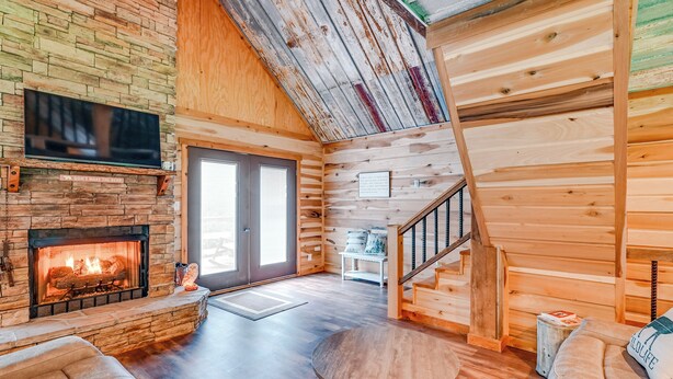 A cozy cabin interior with a stone fireplace, wooden walls, a staircase, and a comfortable seating area. - Kentucky Cabin Retreat w/ Covered Porch & Views (Lewisburg)
