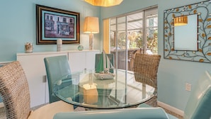 Een eetgedeelte met een glazen tafel, rieten stoelen, een lijstfoto, een lamp en een decoratieve spiegel.