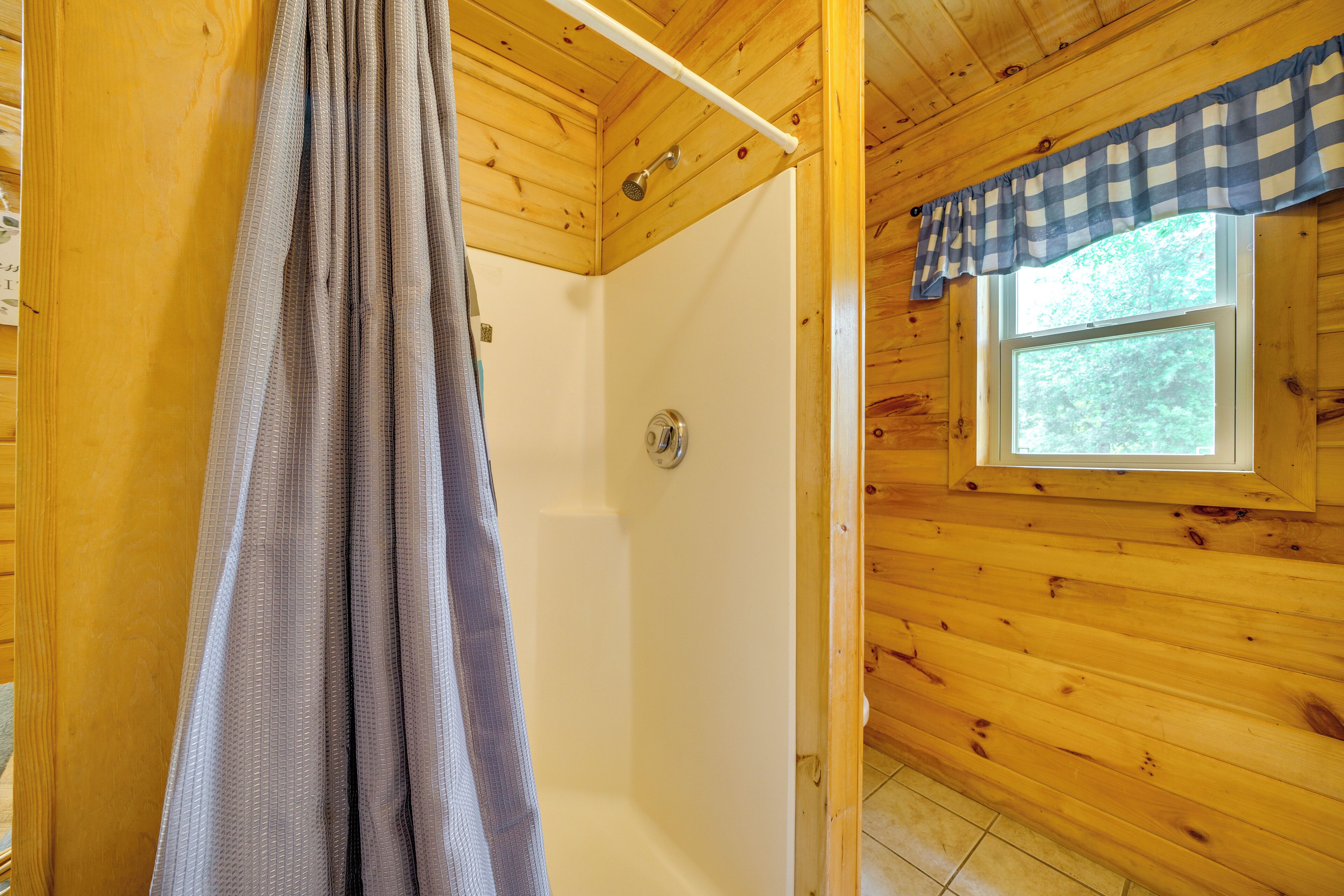 A wooden cabin bathroom with a shower, a window, and a checkered curtain.