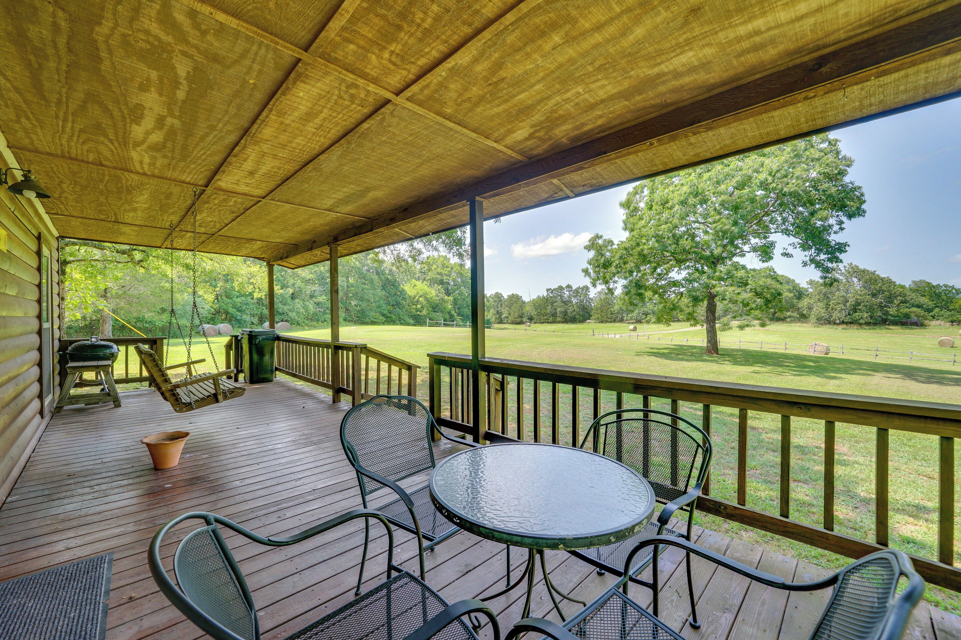 A covered outdoor area with a wooden deck, metal chairs, a table, and a view of a grassy field with trees.