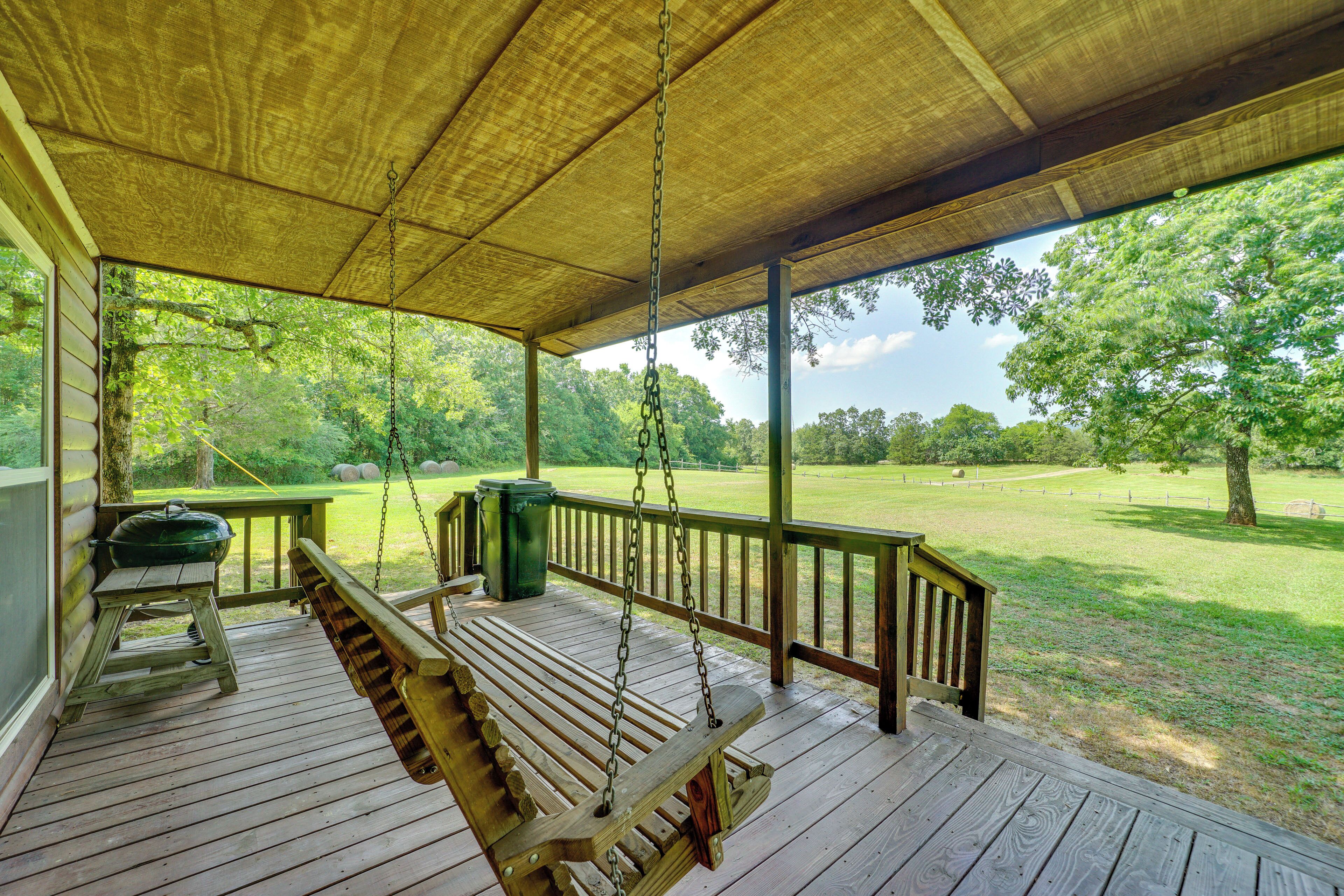 A wooden porch with a swing, a wooden table, and a view of a grassy field.