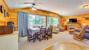 A cozy cabin interior with a dining area, a fireplace, a television, and large windows. - Inlet Lakefront Home: Private Dock & Beach Access! (Inlet)