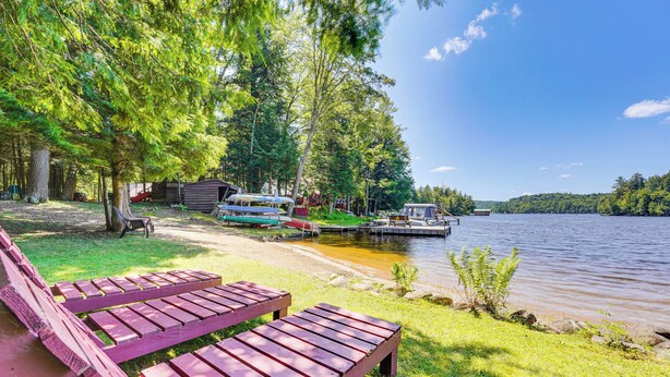 A lakeside picnic area with wooden benches, a dock with boats, and a small cabin. - Inlet Lakefront Home: Private Dock & Beach Access! (Inlet)