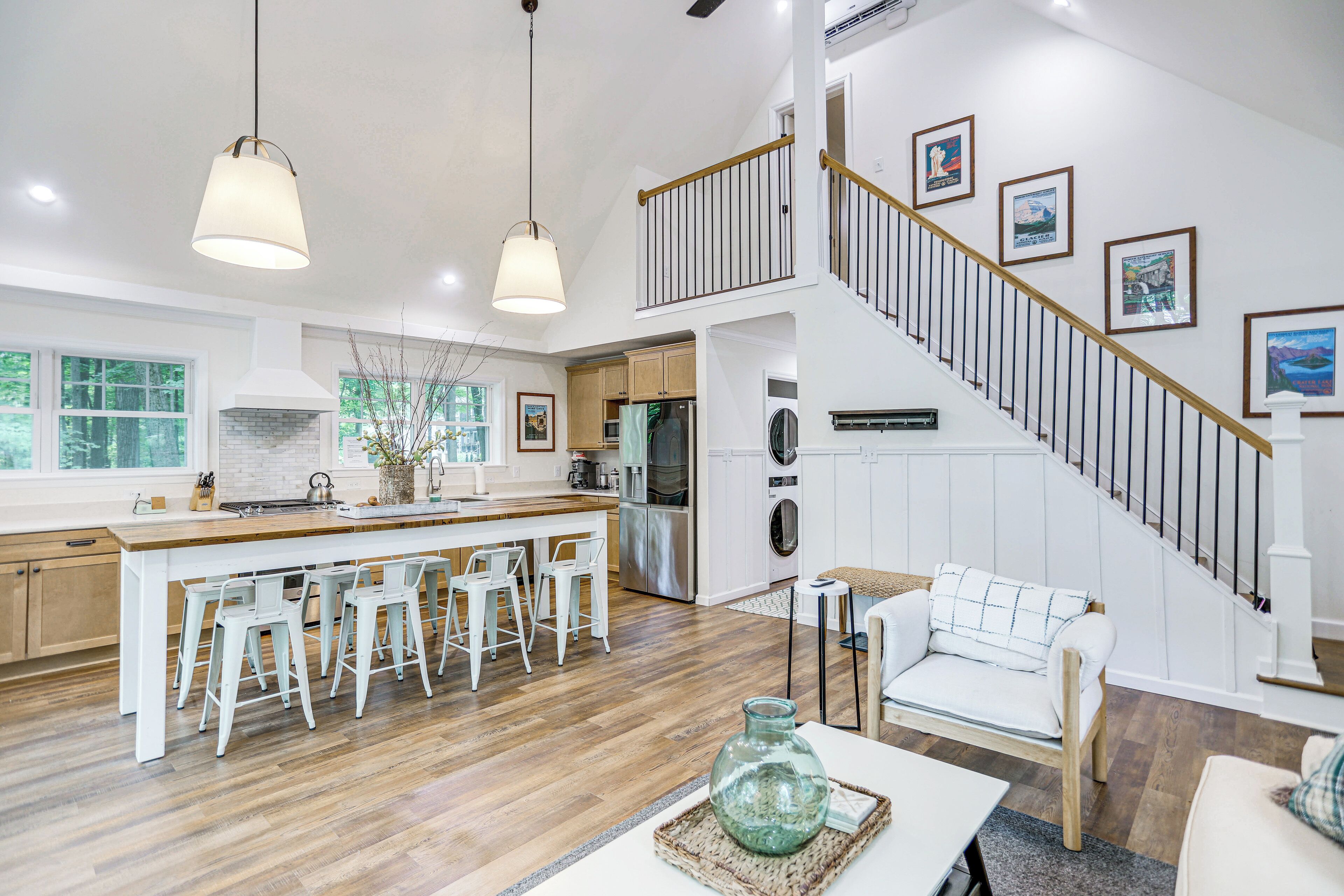 Una cocina amplia con una isla central, taburetes de bar, un comedor con sofá blanco y una escalera decorada con cuadros.